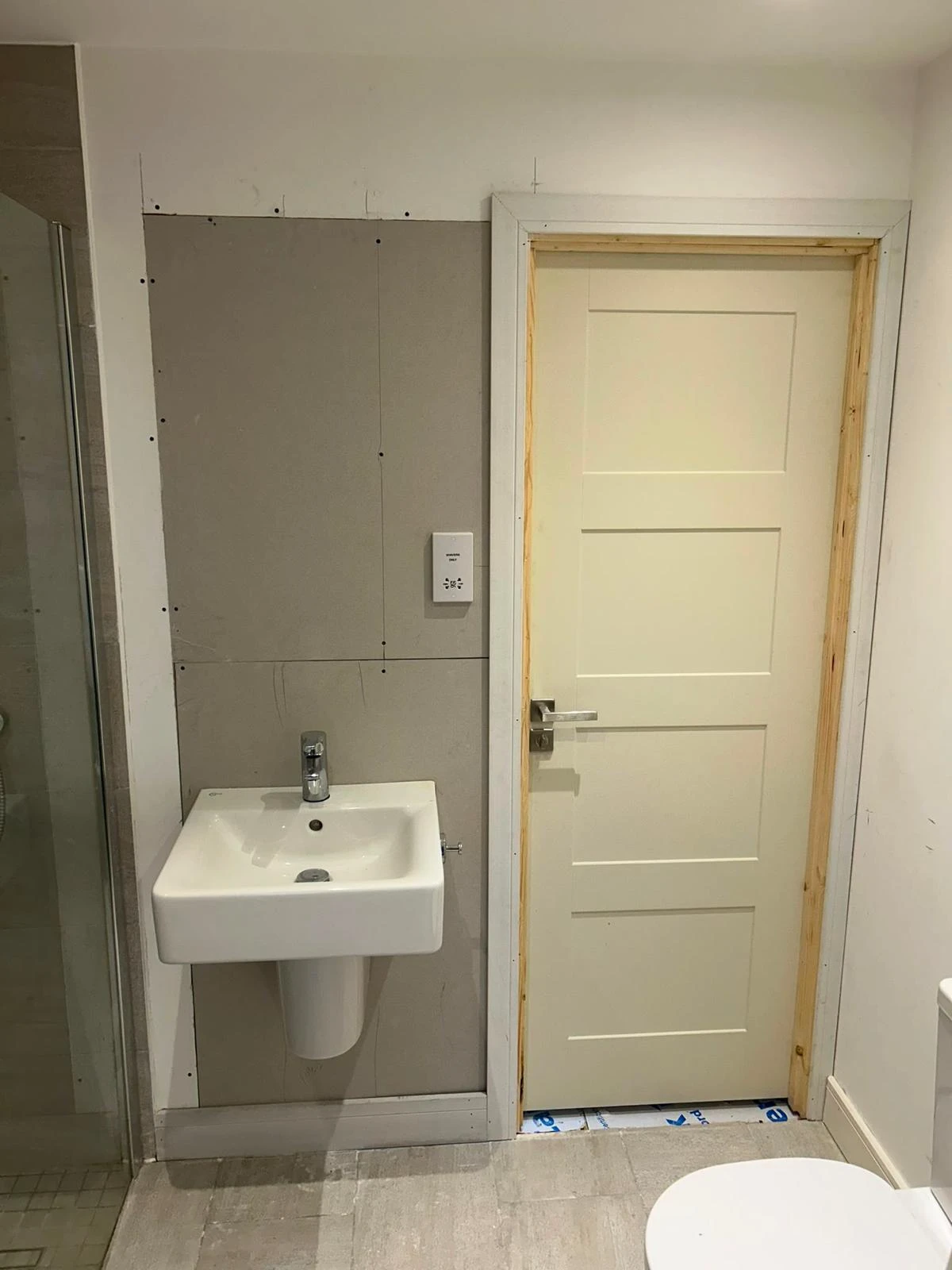 Bathroom with a white wall-mounted sink to the left of a beige five-panel door with an unfinished frame, tile floor, and a shower enclosure partially visible on the far left.
