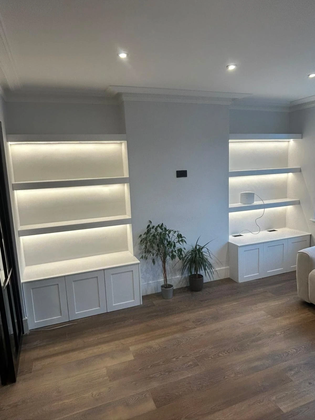 Modern living room corner with two white built-in shelves illuminated by soft LED strip lighting, two potted plants on the floor, and light wood flooring.