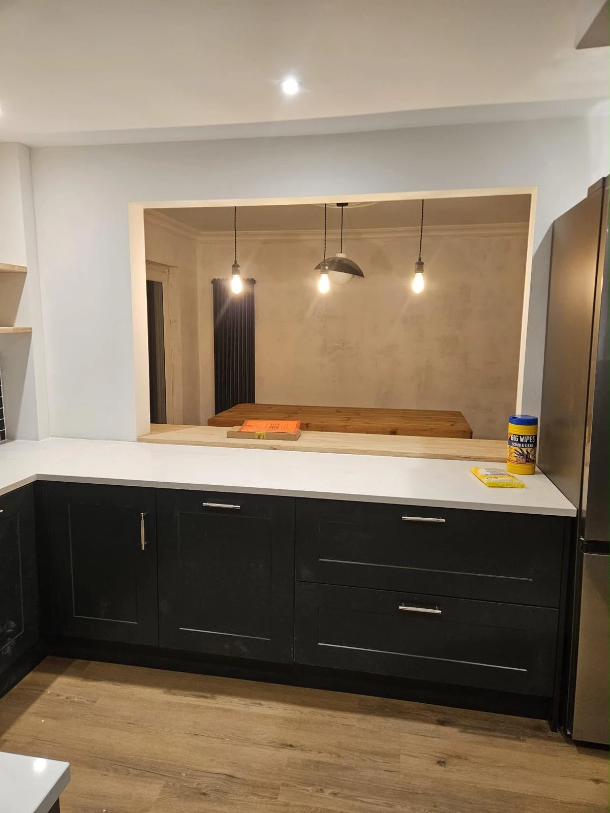 Modern kitchen interior with black cabinets, white countertop, wooden floor, and a pass-through window showing a dining area with three hanging light bulbs and a wooden table.