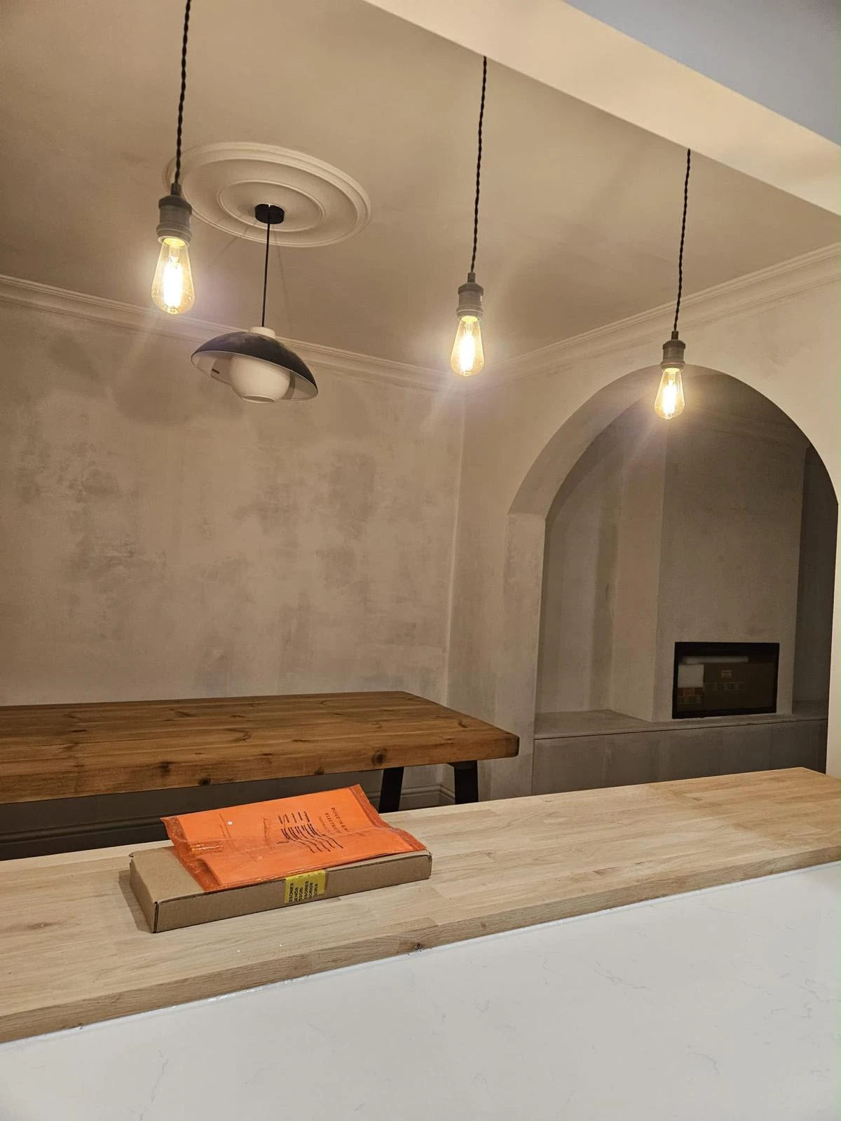 Minimalist kitchen or dining area with three exposed filament pendant lights hanging from the ceiling above a wooden table and a light wood countertop with a small flat cardboard package topped by an orange padded envelope.