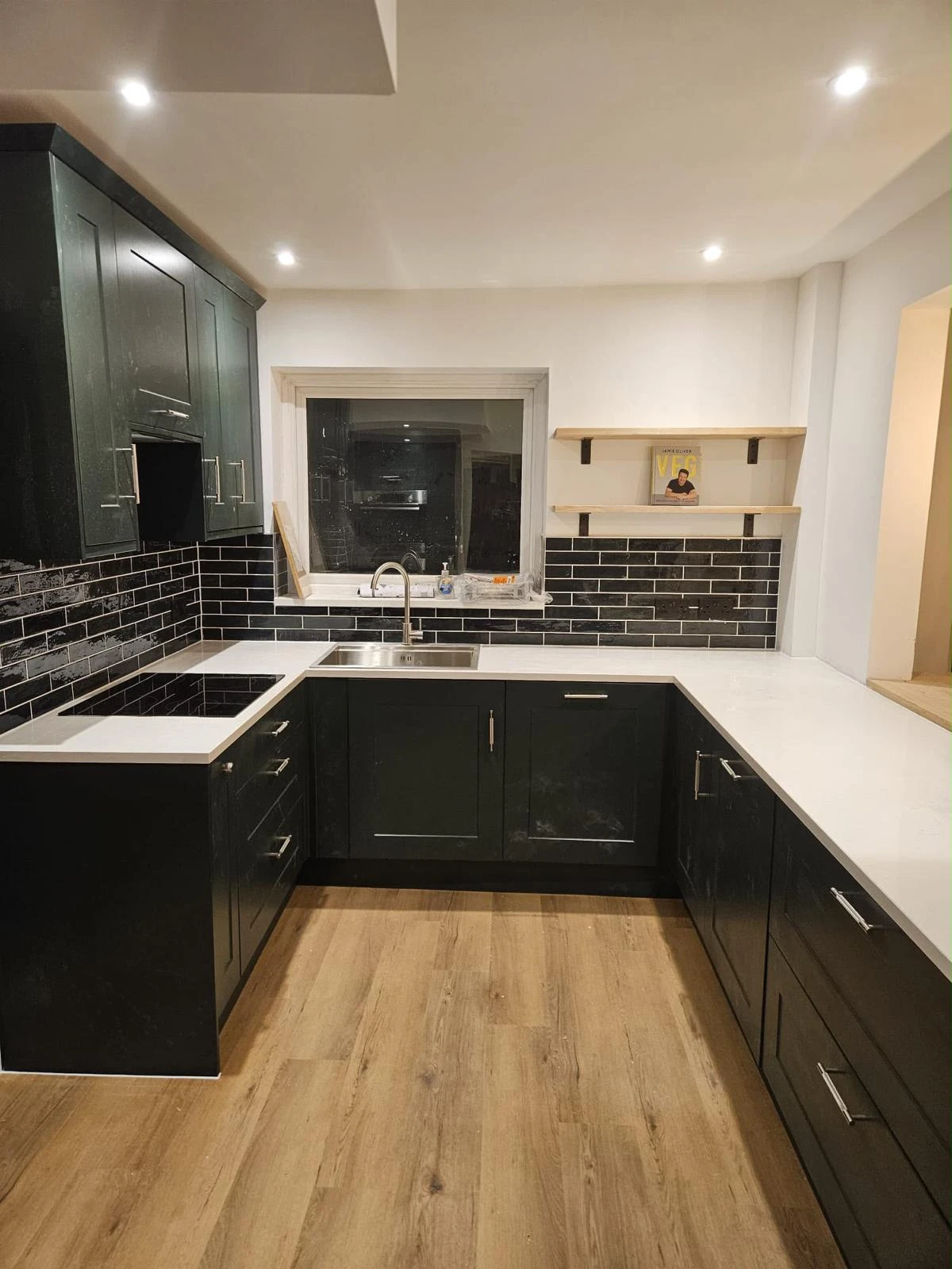 Modern kitchen with black lower and upper cabinets, white countertops, black subway tile backsplash with white grout, stainless steel sink with a chrome faucet, a window above the sink, wooden shelves on the right wall with a kitchen book, and wood-style flooring.