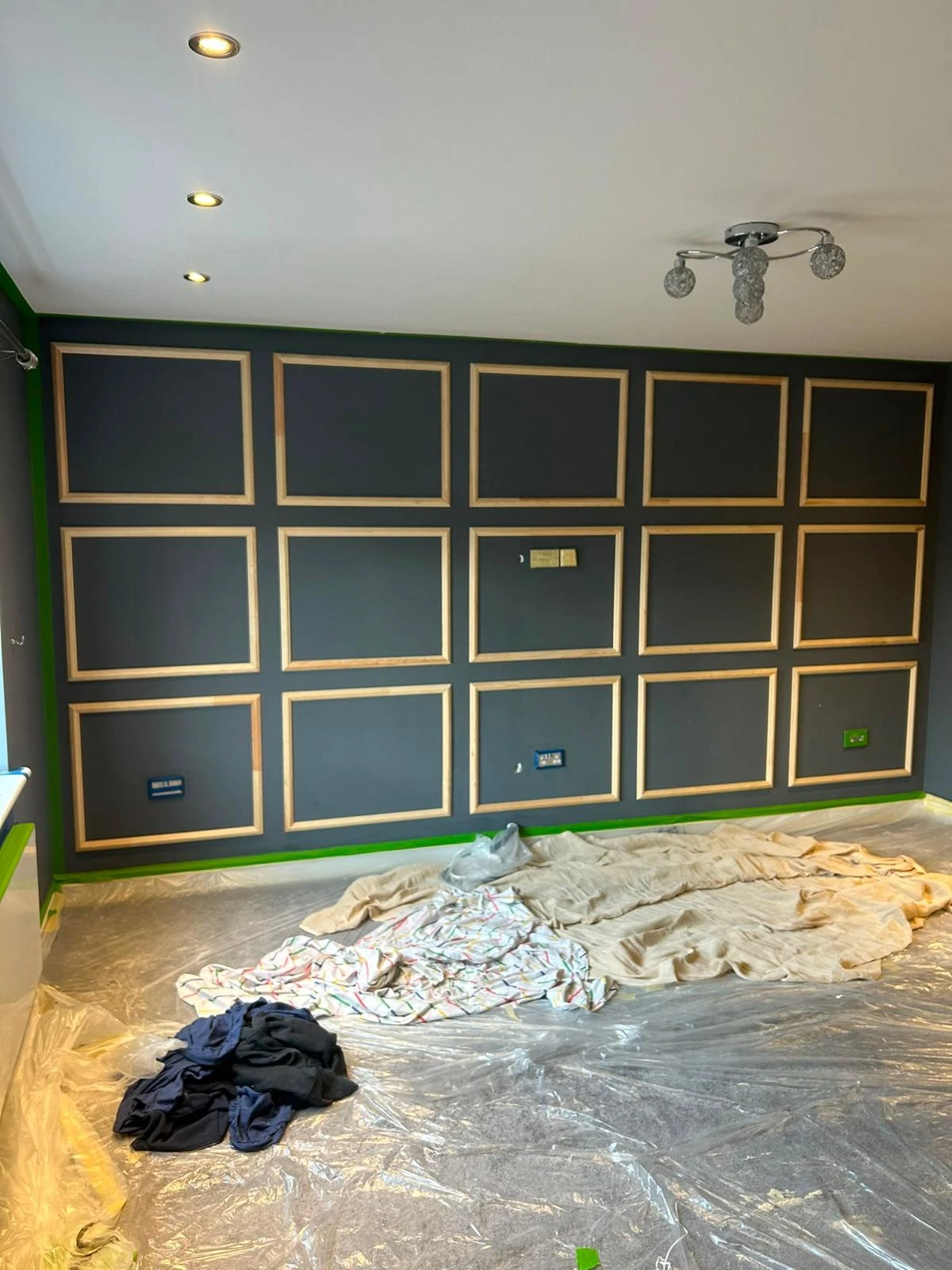 Interior room with a newly painted dark gray accent wall featuring a grid of twelve square wooden trim panels. The floor is covered in clear plastic sheeting and drop cloths, with some clothing or fabric on top. Ceiling has recessed and a decorative light fixture.