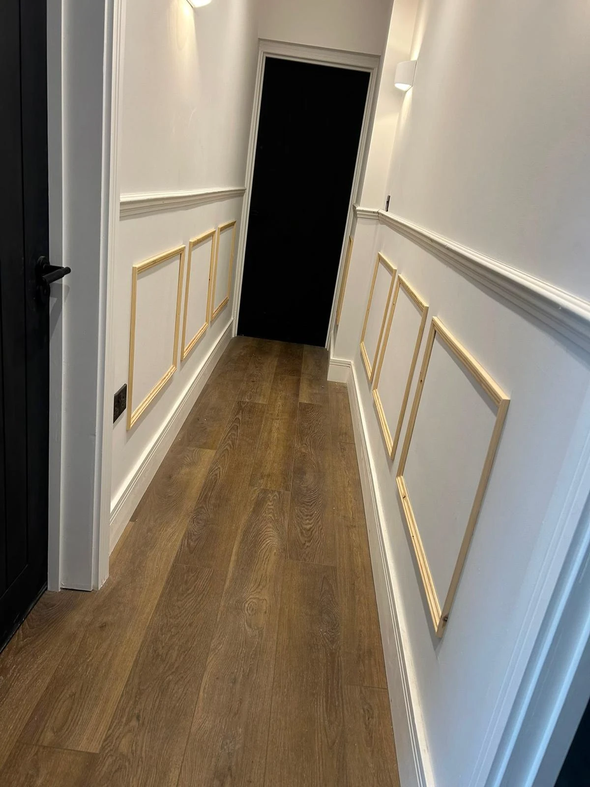 Narrow hallway with wooden floor, white walls featuring decorative wooden frame moldings, black door at the end, and a black door on the left side.