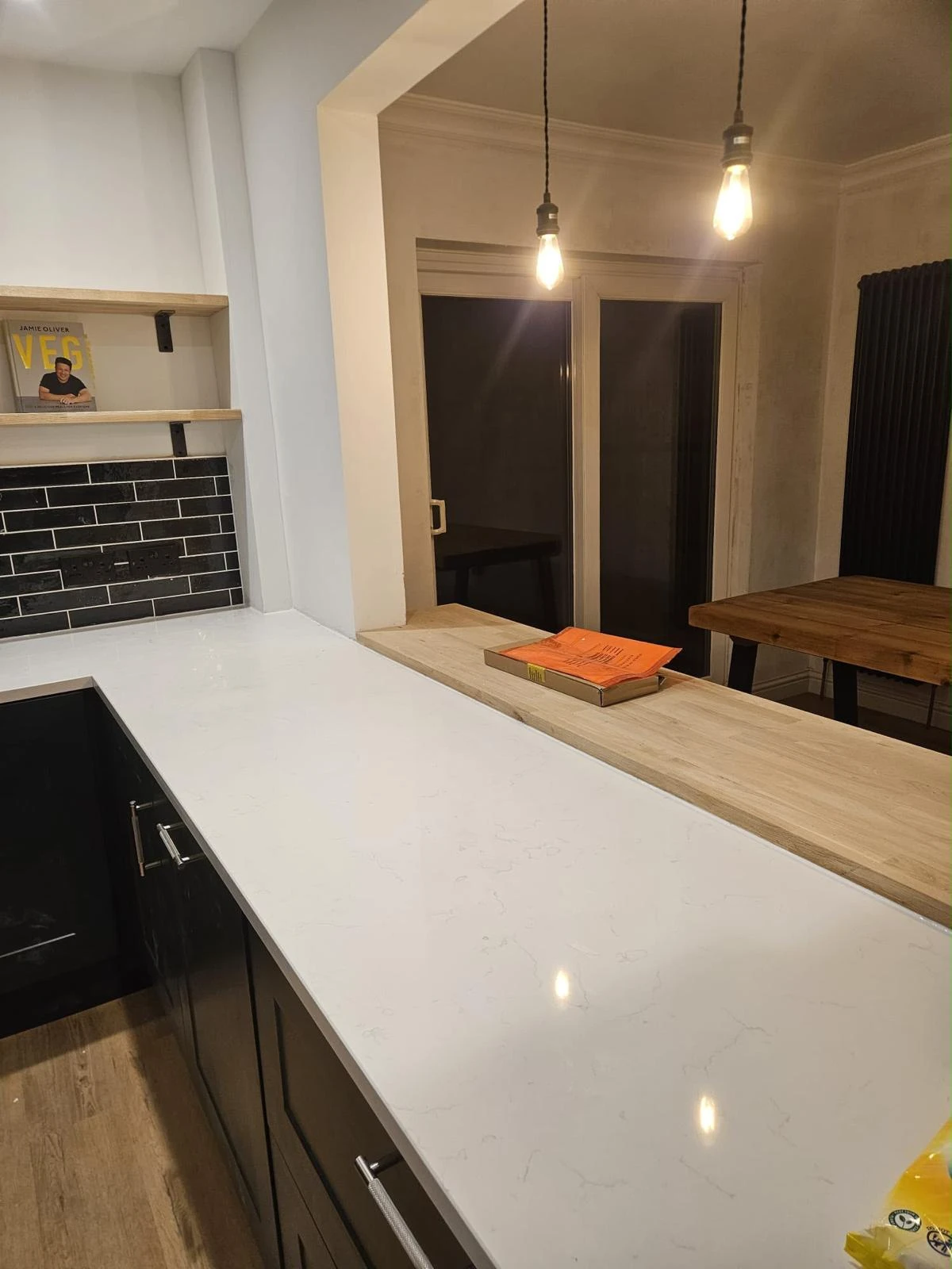 Modern kitchen countertop with white marble surface, black cabinets, and wood accents, featuring two hanging pendant lights and a book on the wooden pass-through ledge.
