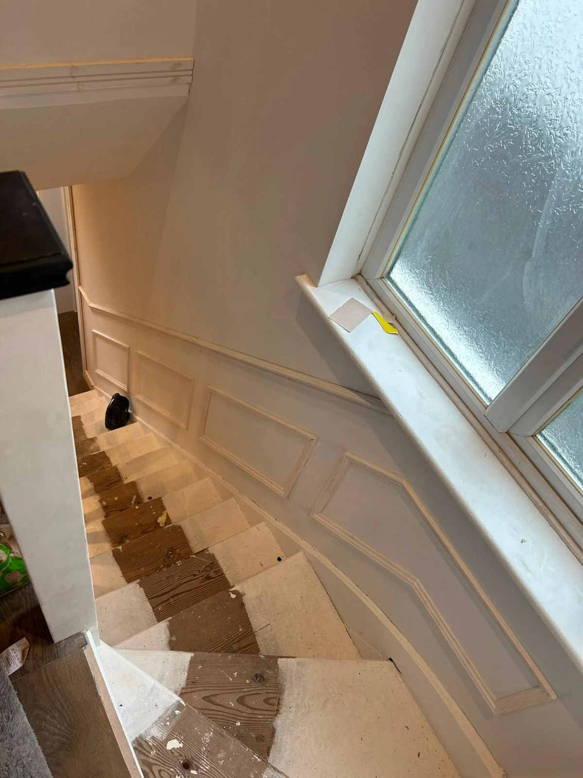 Staircase under renovation with worn wooden steps partially covered by white paint, a textured glass window on the right, and a black object at the bottom of the stairs.