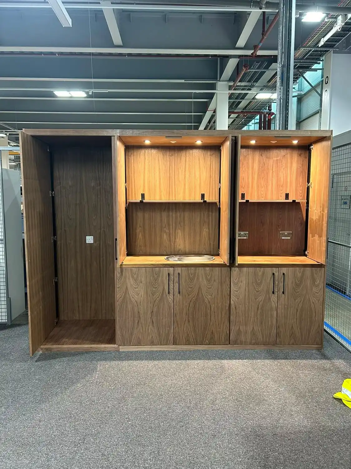 A wooden multi-compartment cabinet unit with three sections; the left section is empty with an open door, the middle section has an open fold-down panel revealing a sink, and the right section has a similar panel with two electrical outlets visible inside, all under small overhead lights.