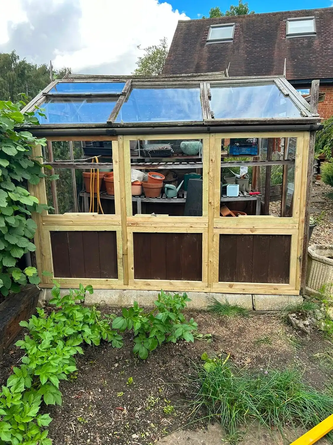 A wooden greenhouse with large glass panels on all sides and roof, containing various gardening pots and tools inside, situated in a garden with green plants and a brick house in the background.