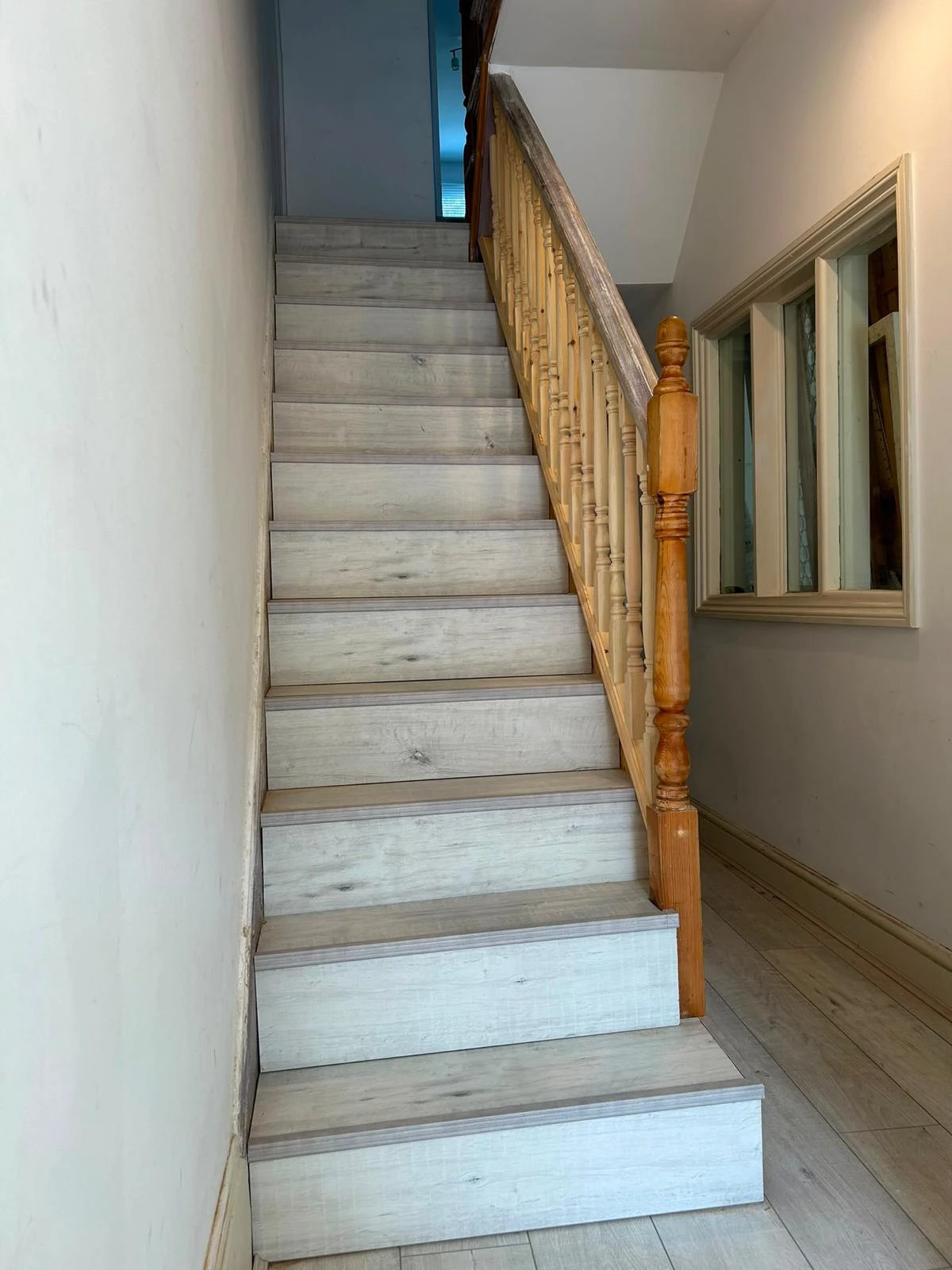 Indoor wooden staircase with light gray steps and a natural wood railing, adjacent to a hallway with a framed window on the right wall.
