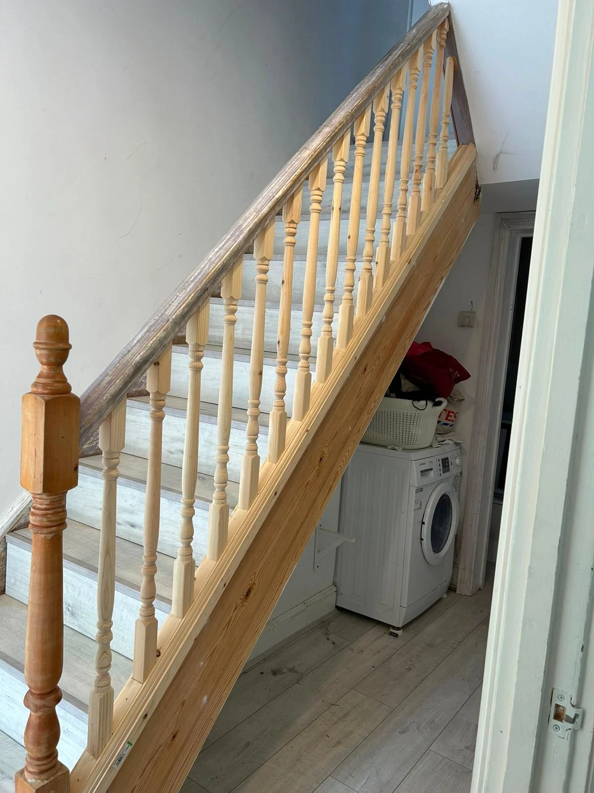Wooden staircase with light-colored steps and natural wood banisters inside a home, with a white washing machine and laundry basket underneath the stairs.