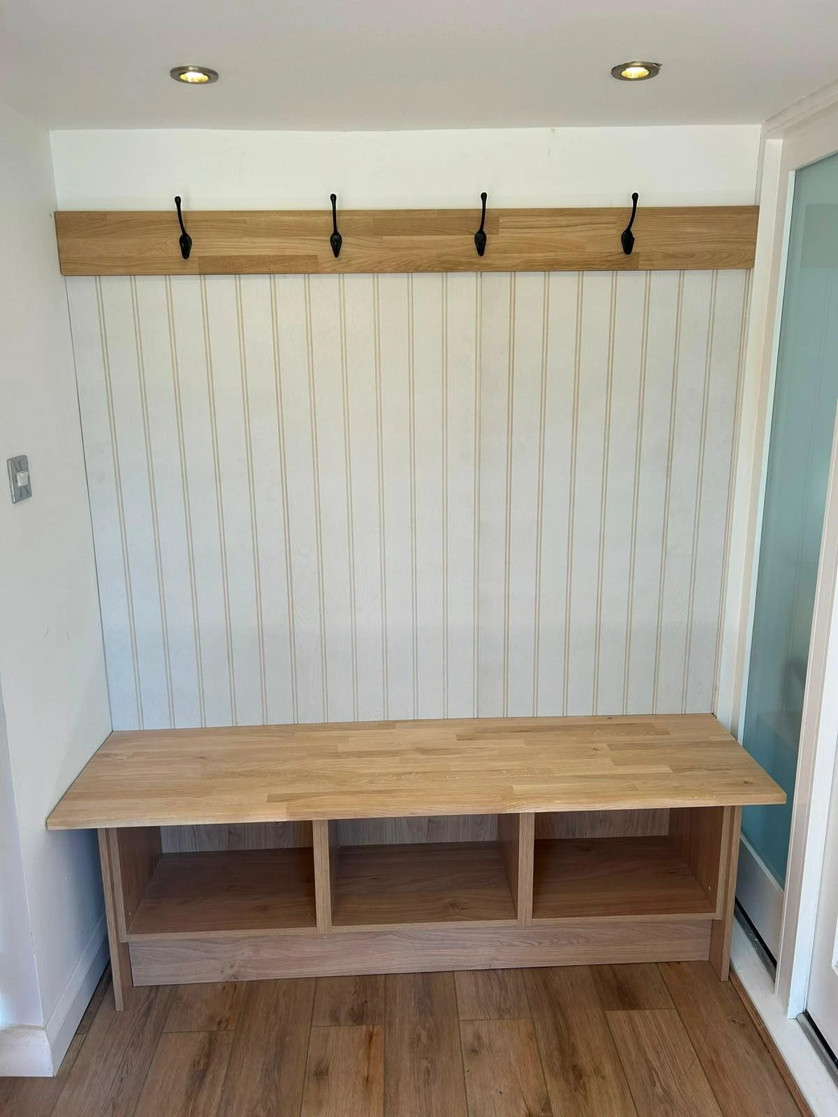 Wooden hall bench with three open cubbies underneath and a back panel with white vertical slats, topped by a wooden coat rack with four black metal hooks, set against a white wall with wood flooring.