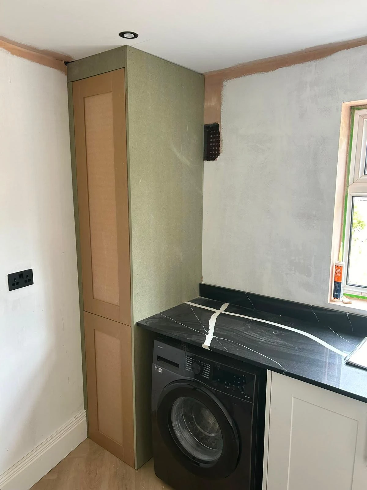 Laundry area with a black front-loading washing machine beneath a black countertop with white veins, adjacent to a tall cabinet and an unfinished wall with a window showing some natural light.