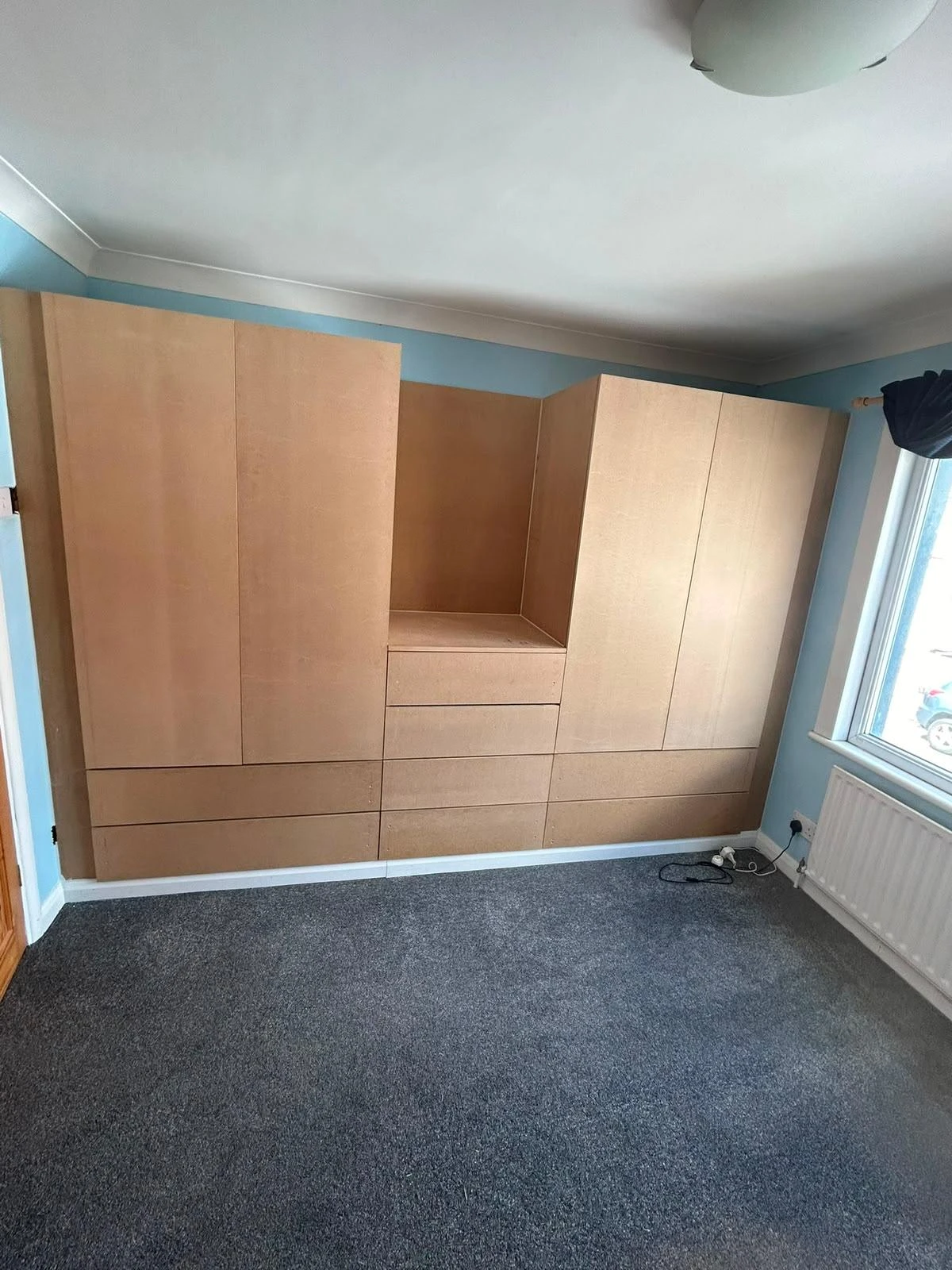 Empty bedroom with installed unfinished light wooden fitted wardrobes covering one wall, a grey carpeted floor, blue walls, a radiator under a window, and a ceiling light.