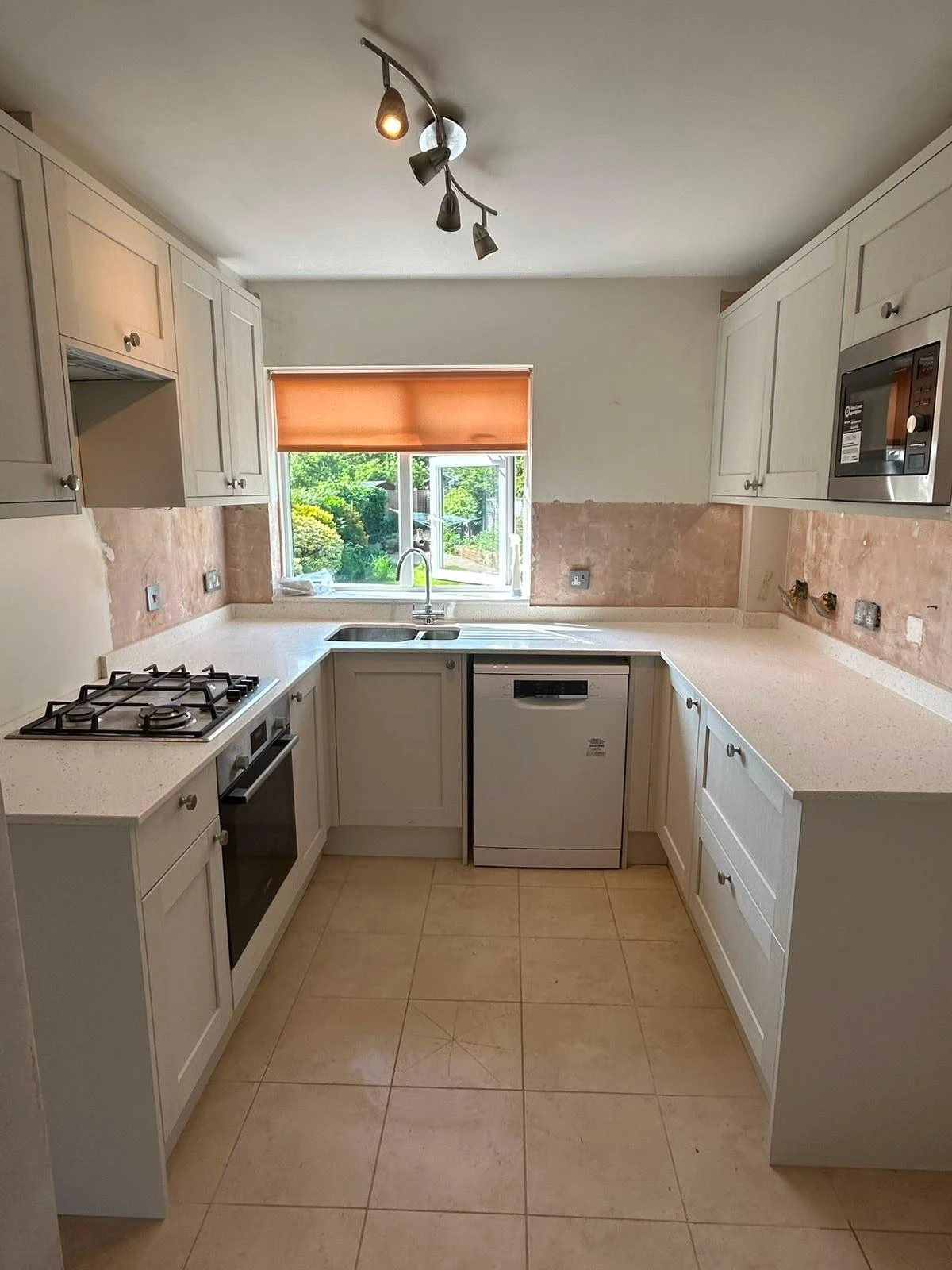 Modern L-shaped kitchen with white cabinetry, white countertops, built-in oven, gas stovetop, dishwasher, double sink under a window with an orange blind, beige tiled floor, and recessed lighting.