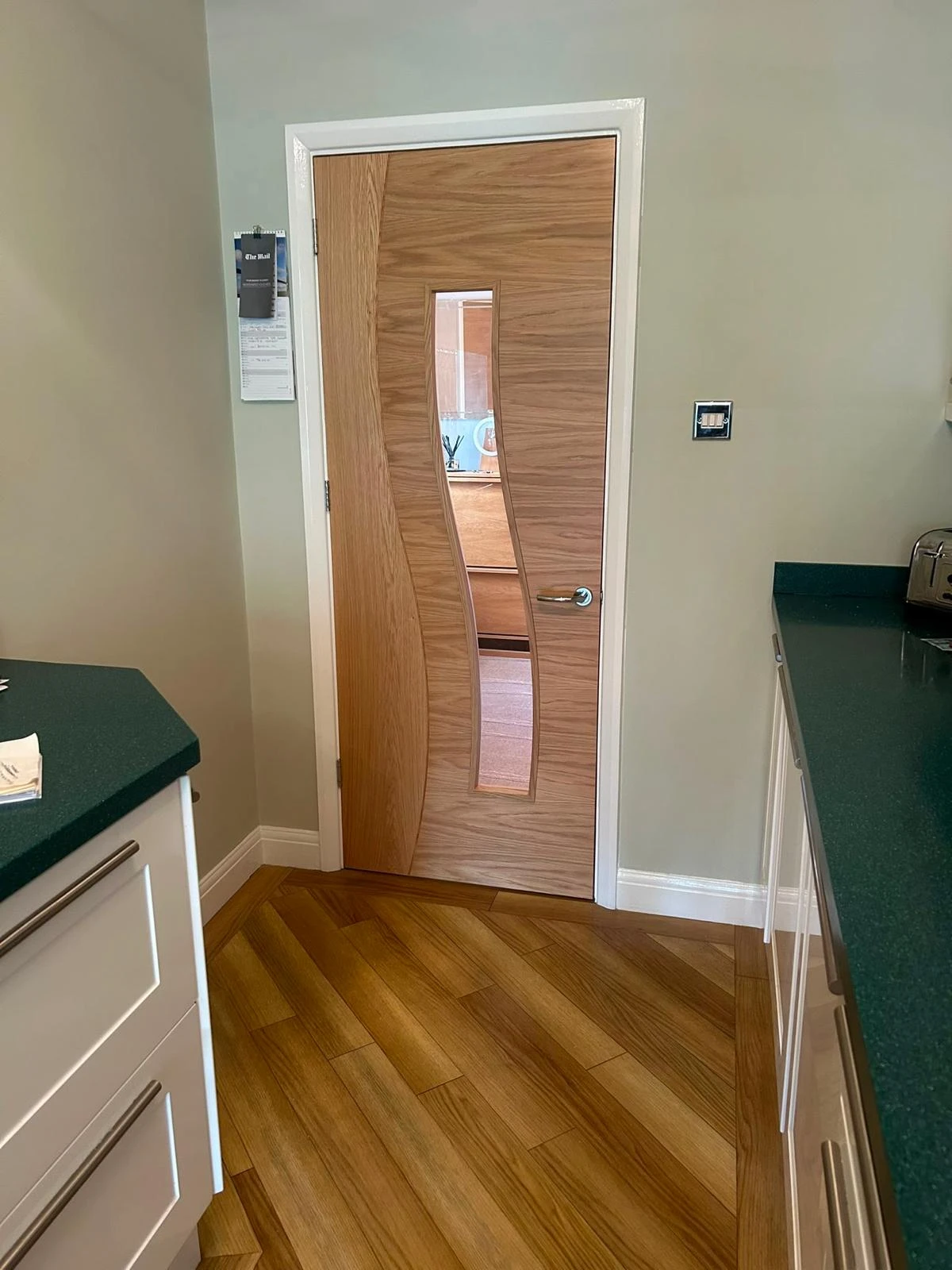 Interior view of a kitchen corner featuring a wooden door with a curved vertical glass panel, green countertops, white cabinetry, and wood-patterned floor tiles arranged diagonally.