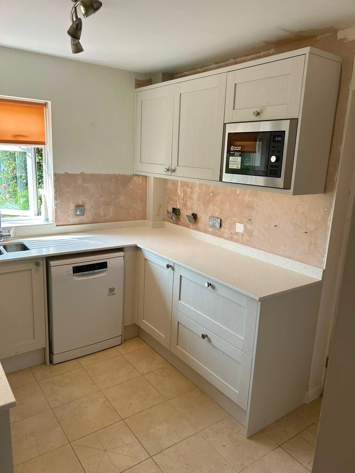 Partial kitchen view showing white cabinets with silver knobs, a built-in microwave, a dishwasher below white speckled countertops, beige tiled floor, and unpainted walls with exposed plaster, next to a window with an orange roller blind.