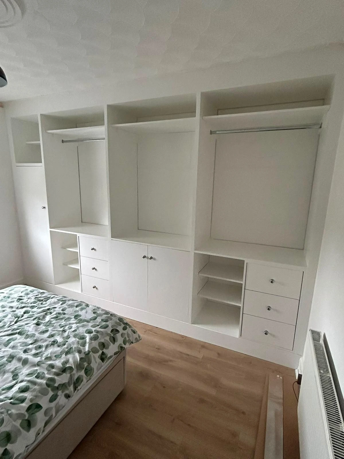 A bedroom corner featuring a large built-in white wardrobe with multiple open shelves, hanging rods, drawers, and closed cabinet doors, next to a bed with a green leaf-patterned duvet cover on a wooden floor.