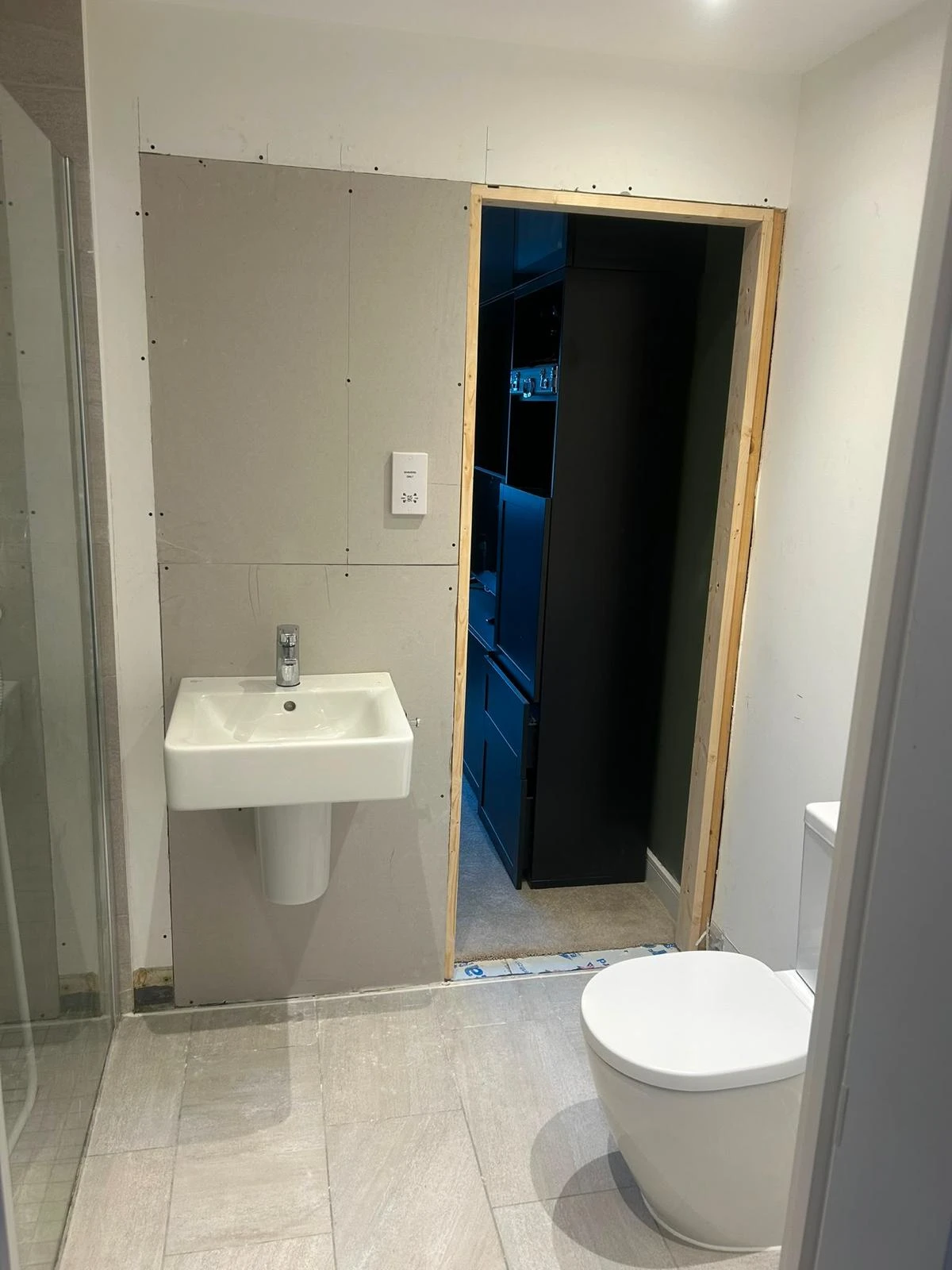 Modern bathroom under renovation with a white wall-mounted sink on the left and a white toilet on the right, an unfinished doorway with visible wood framing leads to a room with dark shelving and carpeted floor.