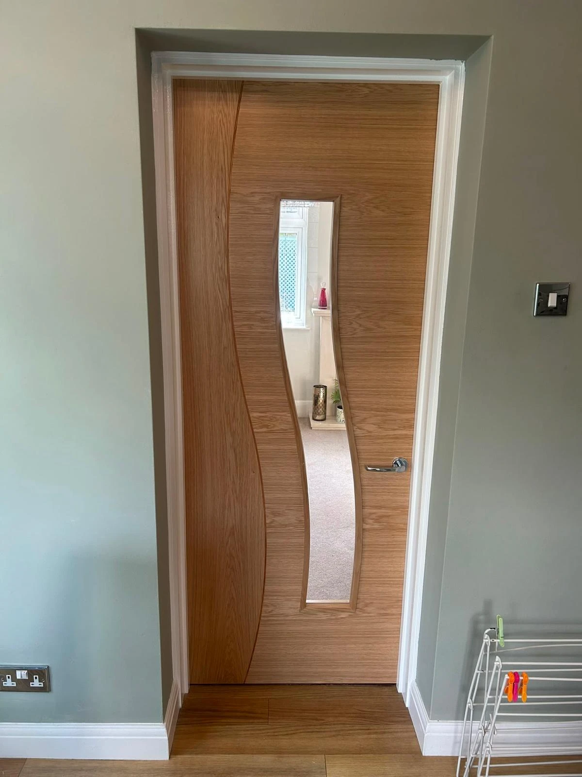 Wooden interior door with a vertical, wavy-shaped narrow mirror panel in the center, framed by white trim, set in a room with light gray walls, wooden floor, a white electrical outlet on the left wall, light switch on the right, and a white drying rack with colorful clips in the bottom right corner.