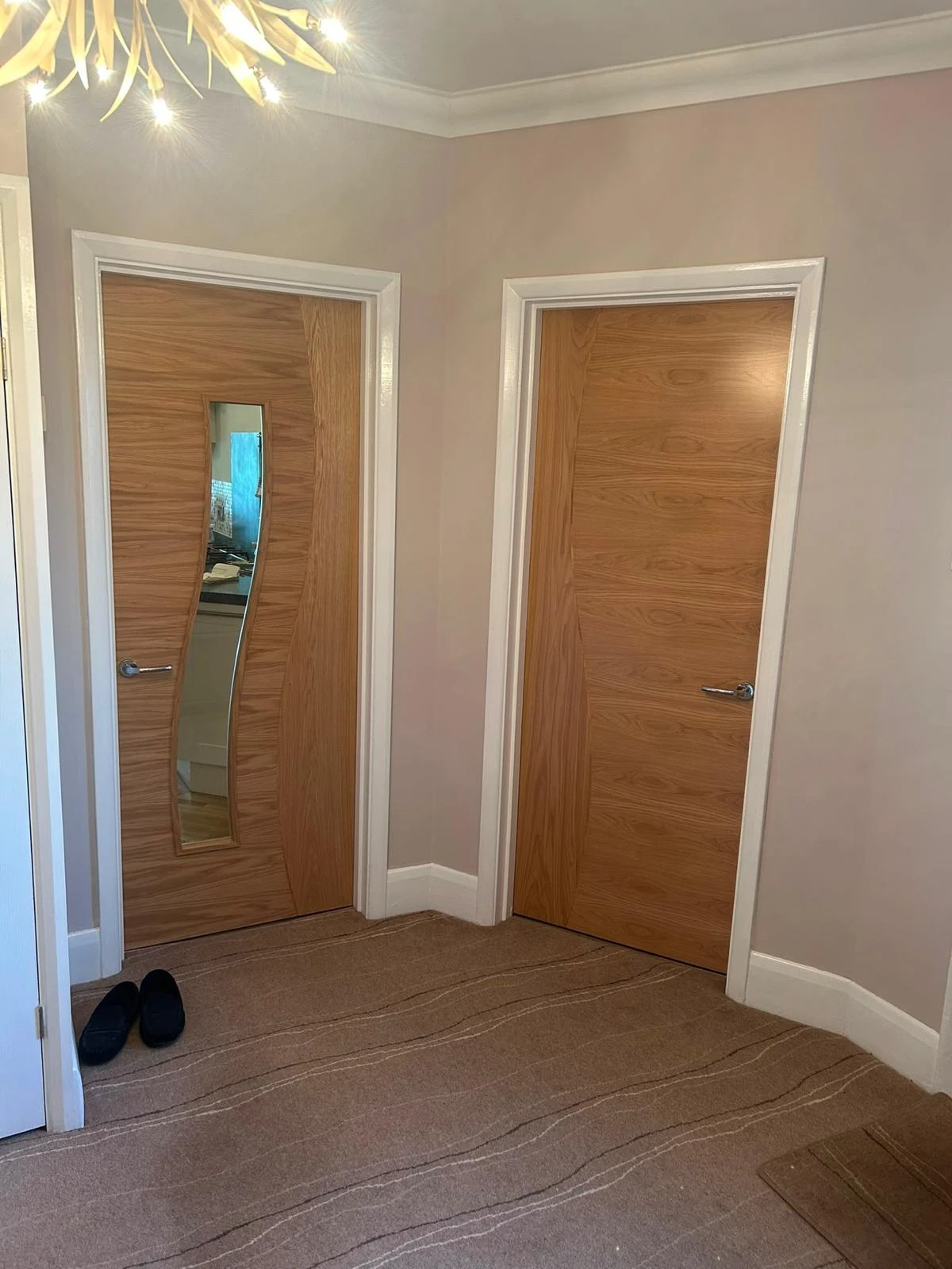 Interior corner of a room with two wooden doors featuring modern wavy patterns; the left door has a narrow vertical mirror inset and silver handle, and the right door has a silver handle. Beige carpet with subtle wavy lines covers the floor, and a pair of black slippers is placed near the left door. The walls are painted light beige, and part of a white door frame is visible on the far left. A modern ceiling light fixture with multiple bulbs is partially visible at the top left corner.