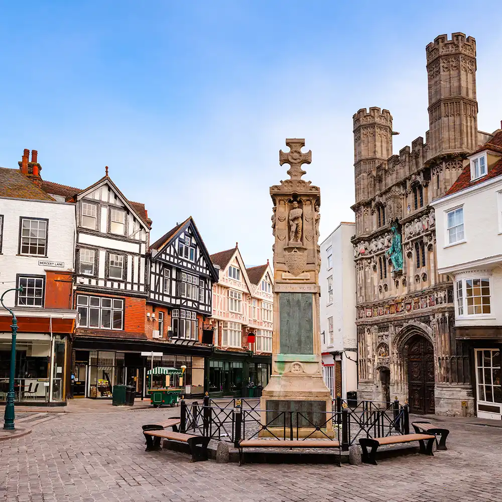 Historic town square with a stone war memorial monument featuring carved figures and a cross on top, surrounded by wooden benches and wrought iron fence; traditional half-timbered and brick buildings and a large ornate stone building with tall towers in the background under a clear blue sky.