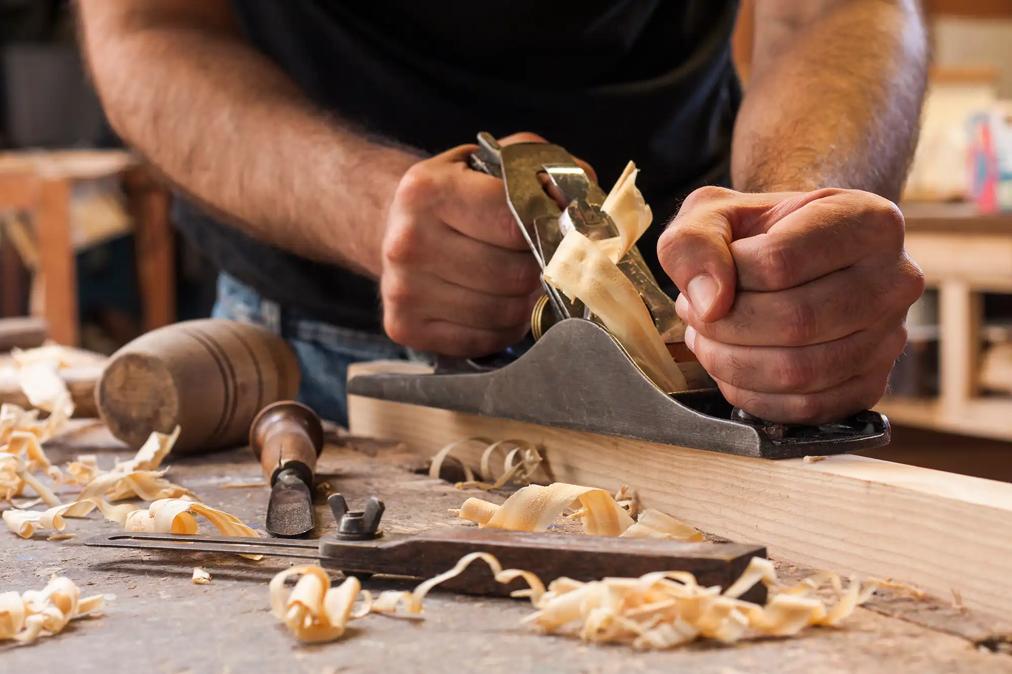 Close-up of a person using a hand plane to shave thin curls of wood from a wooden plank on a workbench, surrounded by wood shavings and woodworking tools.
