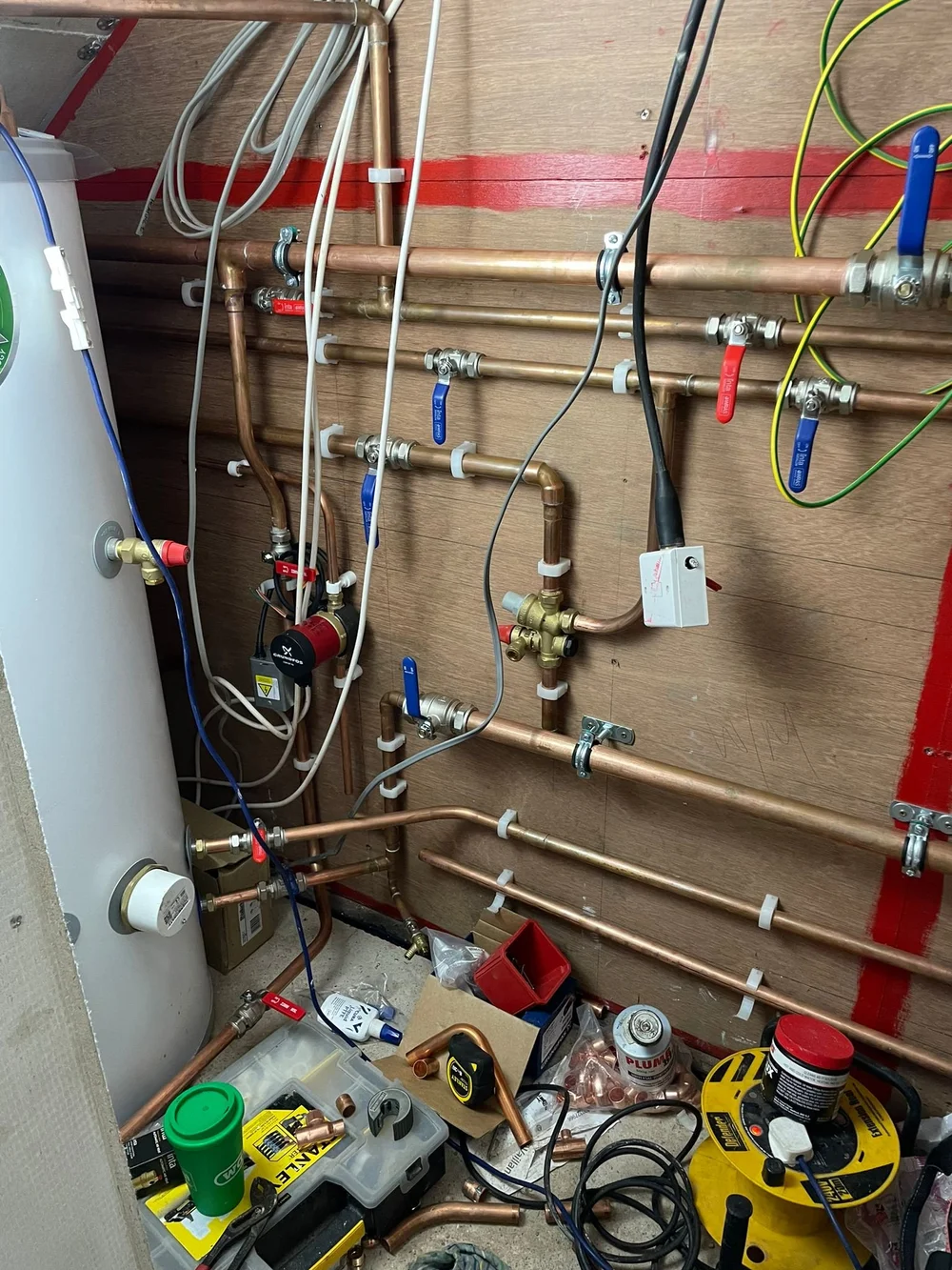 Copper plumbing pipes with valves mounted on wood wall and plumbing tools on floor in utility room.