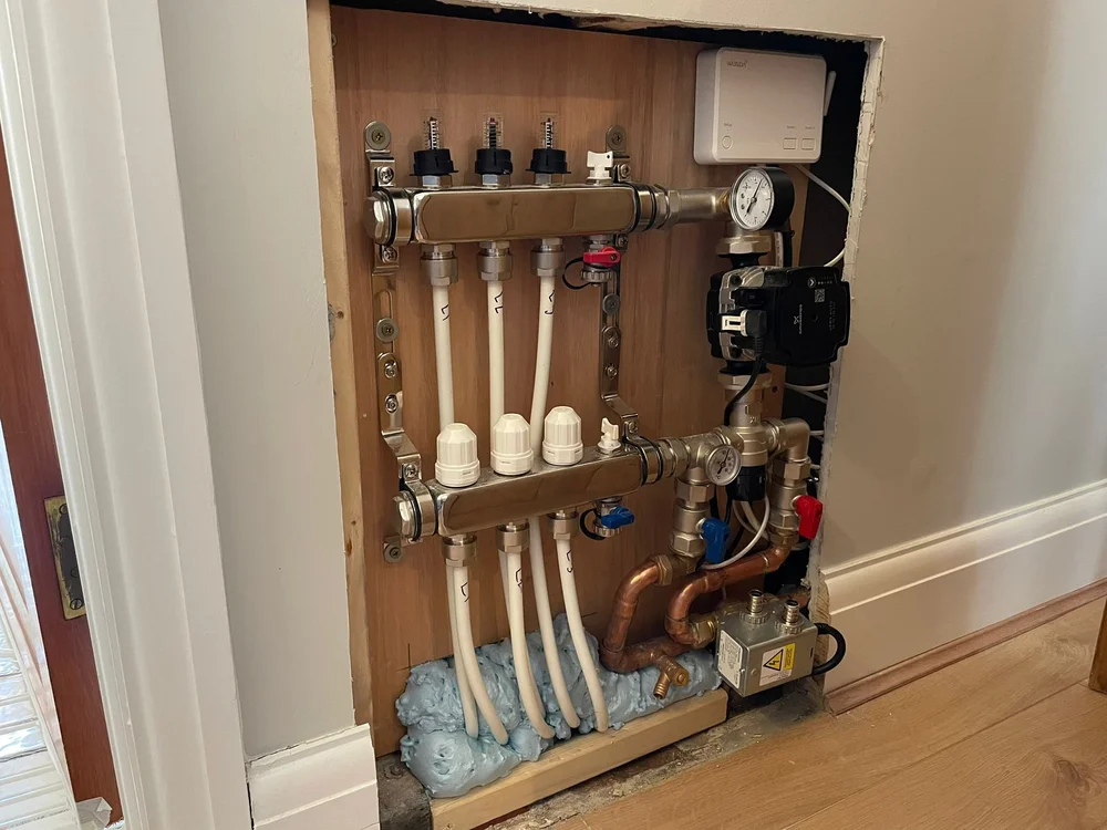 Wall-mounted plumbing manifold with pipes, valves, gauges, and insulating foam at bottom in a wood panel cutout.