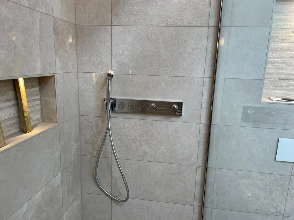 Modern shower wall with chrome handheld shower head, knobs, and inset shelf with gold decorative elements.