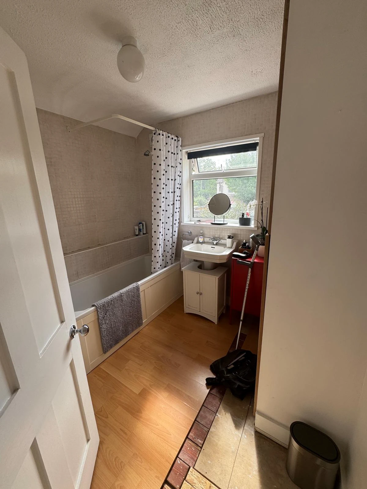A well-lit bathroom with a bathtub, sink, and window. Notable features include a shower curtain, wooden floor, and a waste bin.