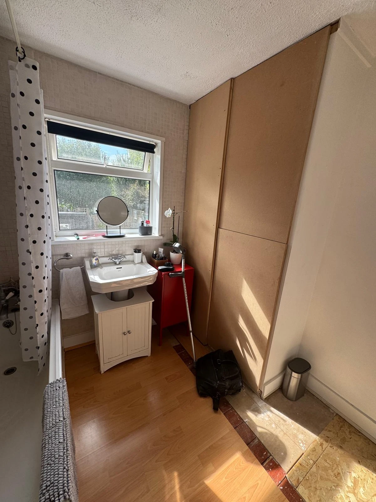 Bright bathroom with a window, sink, and mirror; features a tub, red cabinet, and a dark bag on the floor.
