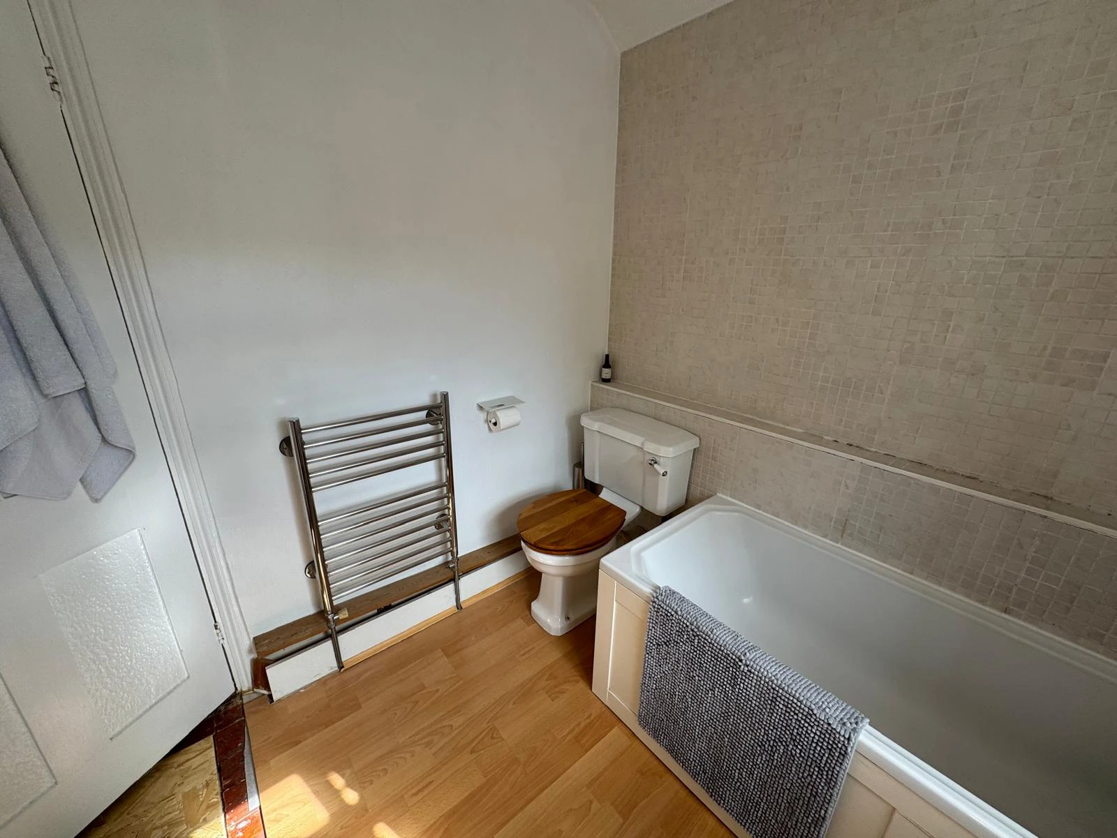 Modern bathroom with a bathtub, toilet, heated towel rail, wooden accents, and light-colored tiled walls. Bright and clean ambiance.