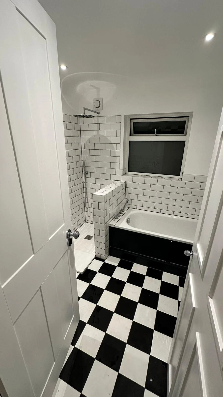 Modern bathroom with a black and white checkered floor, white tiled walls, bath, and shower area. Bright overhead lighting.