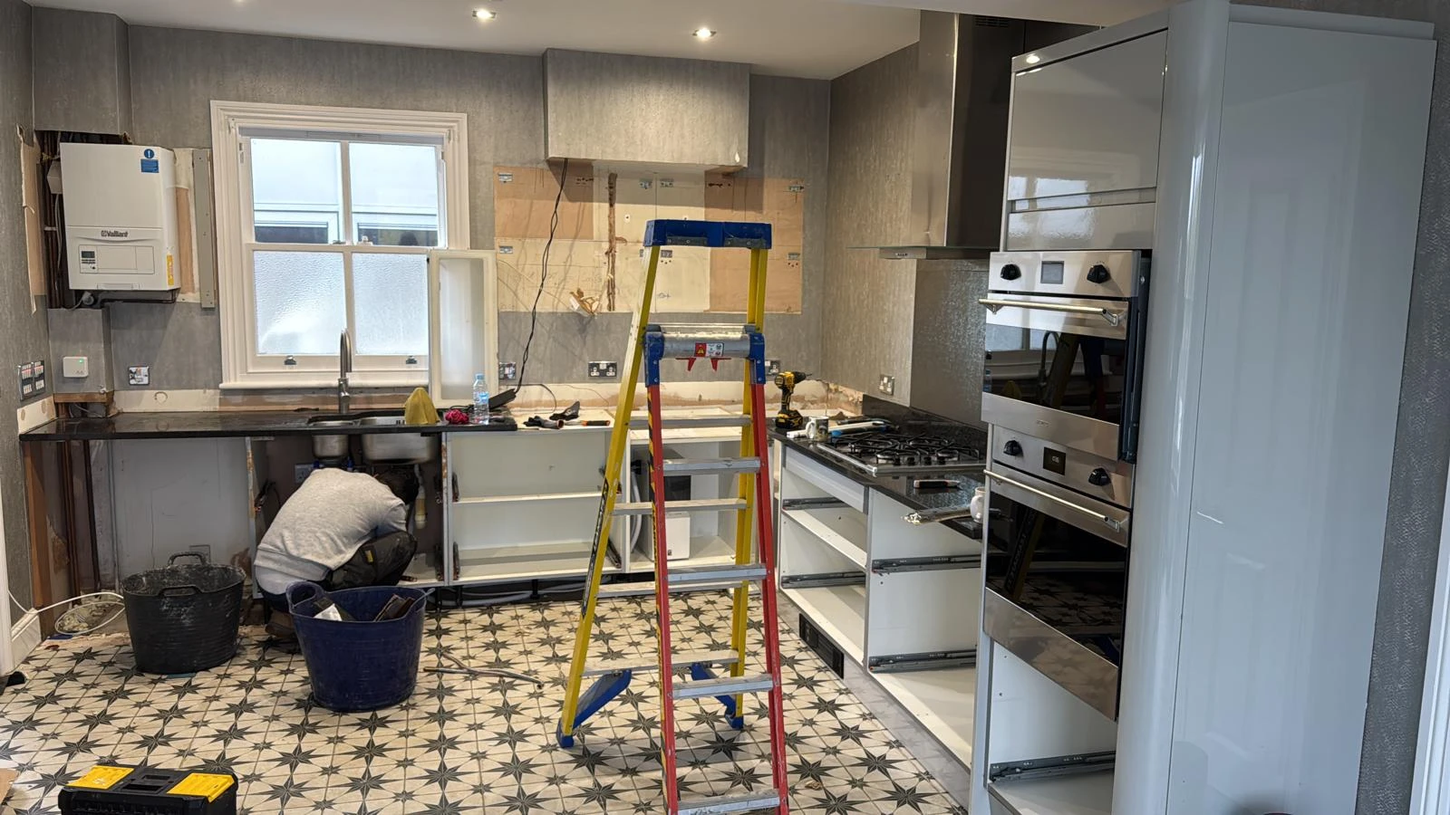 A partially renovated kitchen with a worker crouching under the sink, a ladder, appliances, and patterned flooring visible.