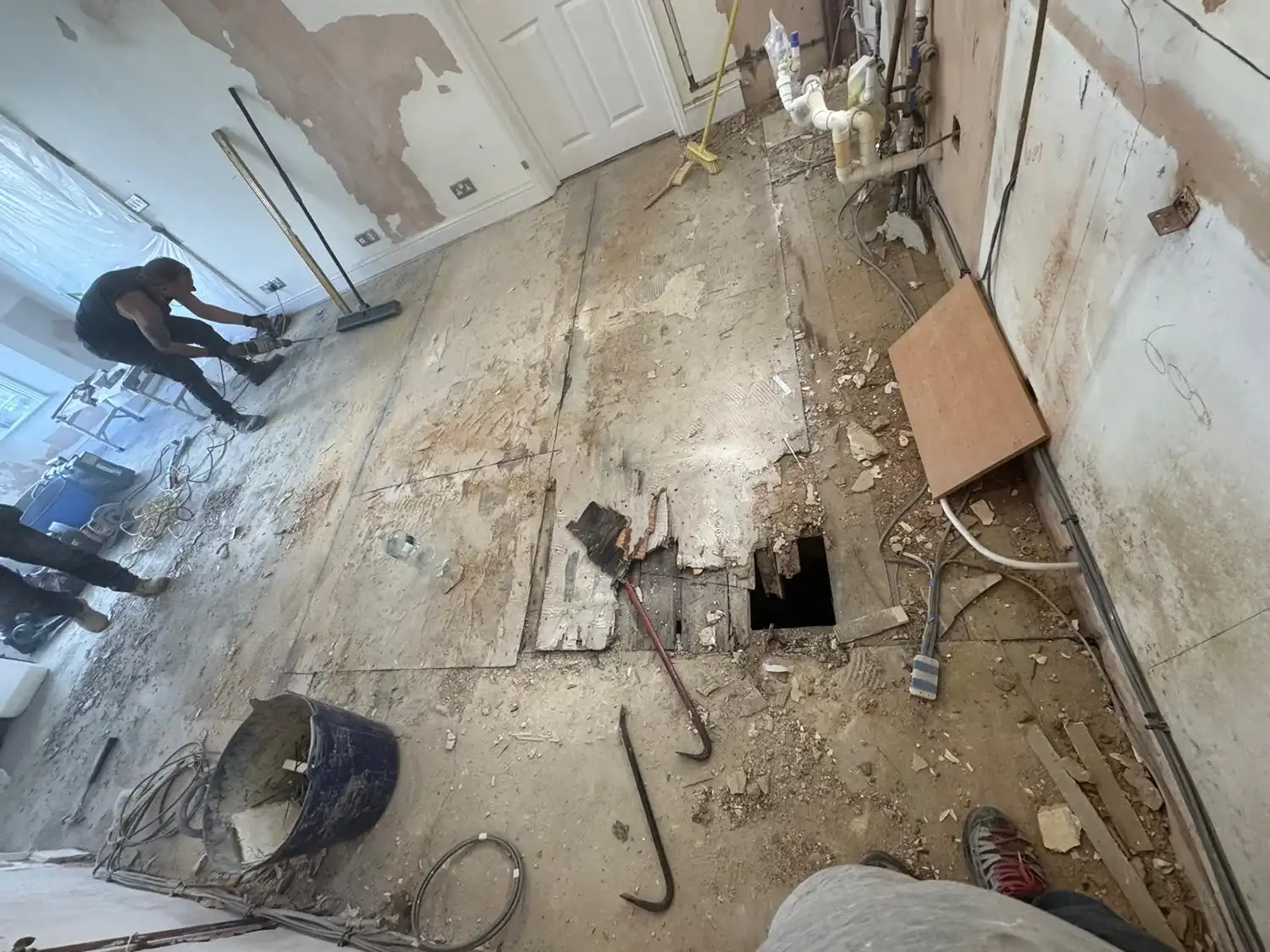 A partially demoed room with exposed floor, debris, and tools. Two workers are visible, one using a saw near a hole in the floor.