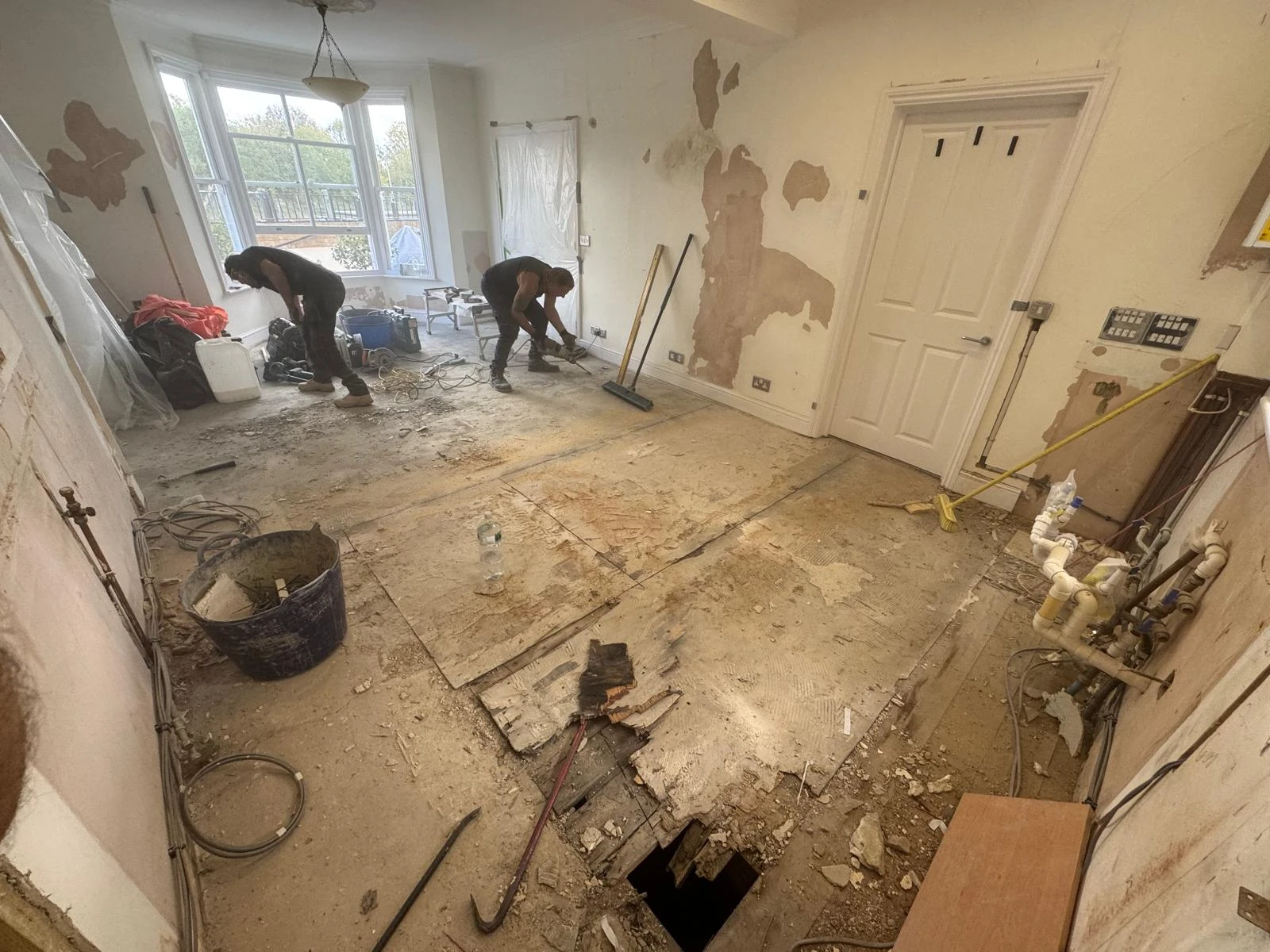 Workers renovate a room with exposed floors and walls. Tools and debris are scattered around. Natural light enters through large windows.