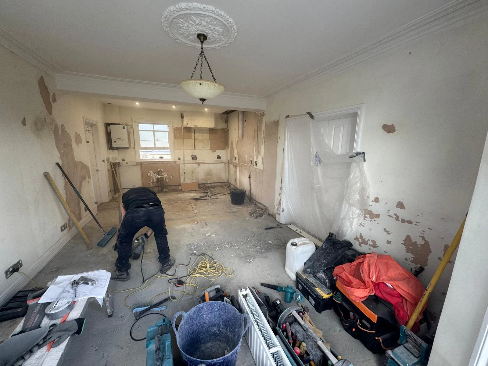 An unfinished room with stripped walls and cluttered floors. A person bends down working, surrounded by tools and construction debris.
