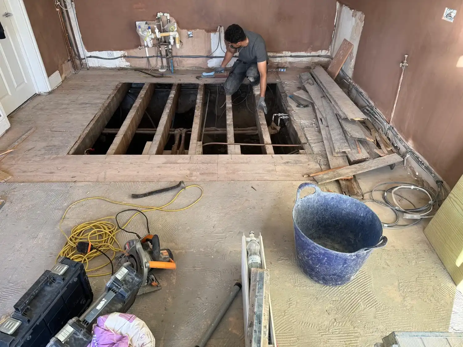 A person kneels on a partially removed wooden floor, revealing a space beneath. Tools and materials are scattered around the room.