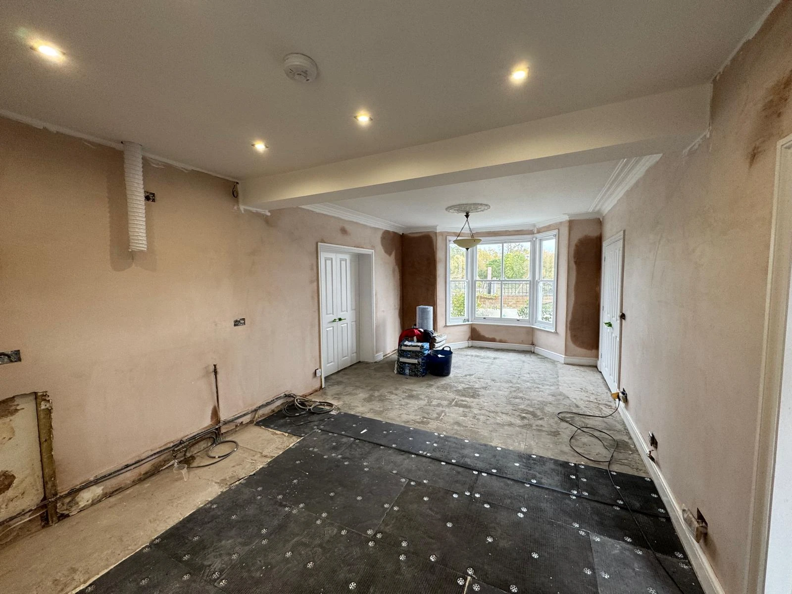 Renovated room with bare walls, unfinished flooring, and a large window. Tools and materials are scattered around, indicating ongoing work.