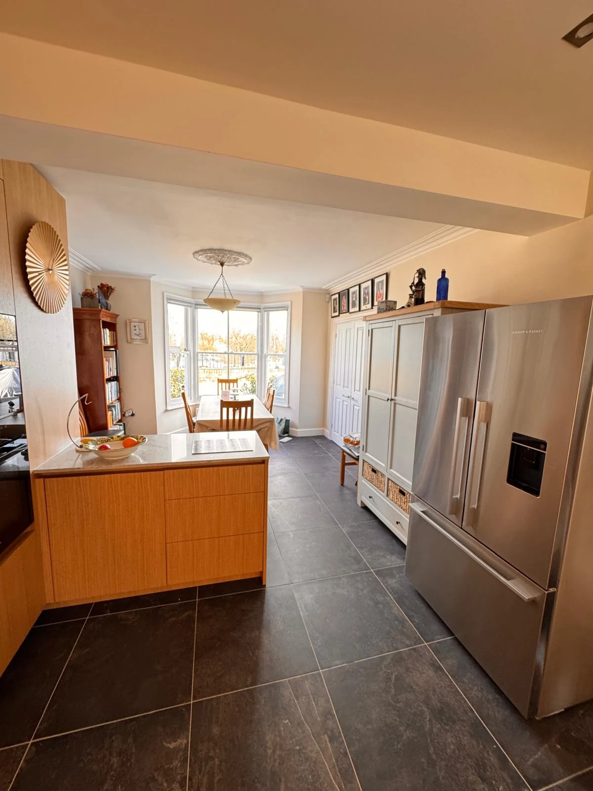 Modern kitchen with light wood cabinets, stainless steel refrigerator, and a dining area with a bay window and artwork on the walls.