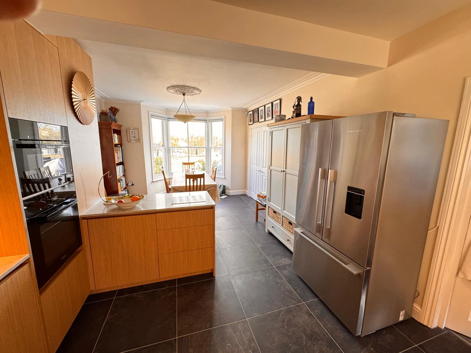 Modern kitchen with stainless steel refrigerator, wooden cabinets, dining area, and large windows providing natural light.