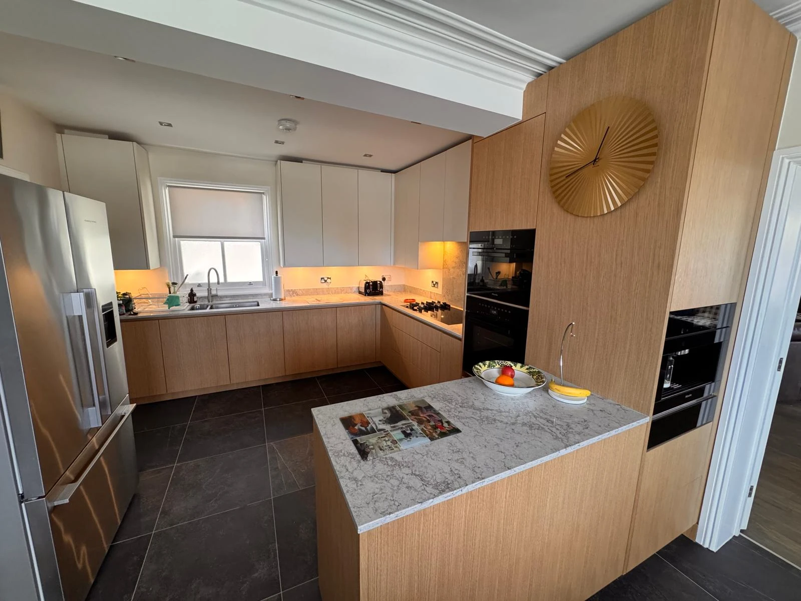 Modern kitchen with wood cabinetry, stainless steel fridge, stone countertop, and decorative fruit bowl. Bright, spacious, and stylish design.