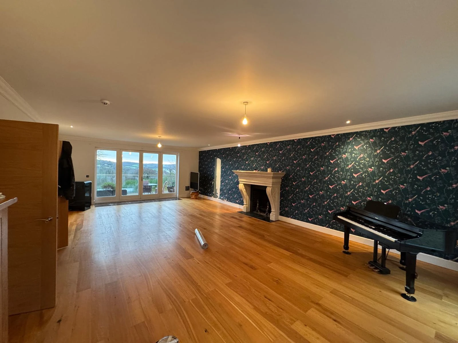 Spacious living room with wooden flooring, a decorative fireplace, and a piano against a floral wallpaper backdrop. Bright natural light from large windows.
