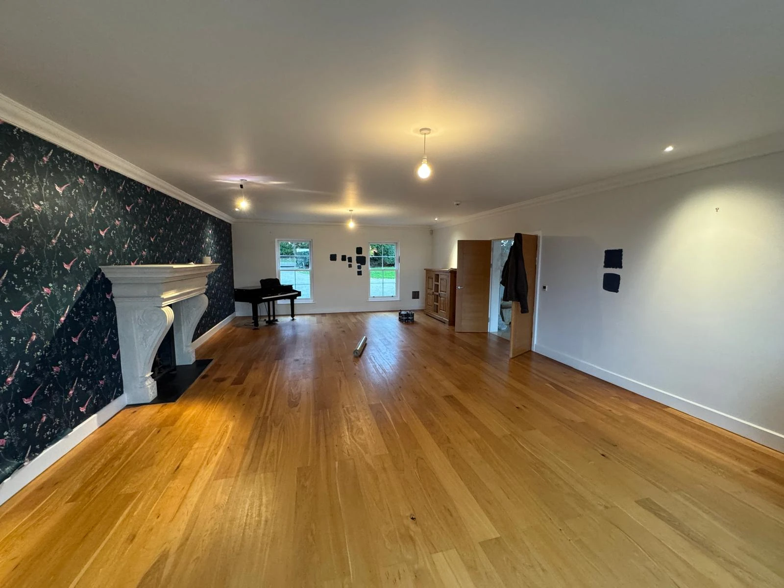 Spacious living room with wooden flooring, a floral wallpapered wall, a fireplace, and a piano. Natural light from large windows.