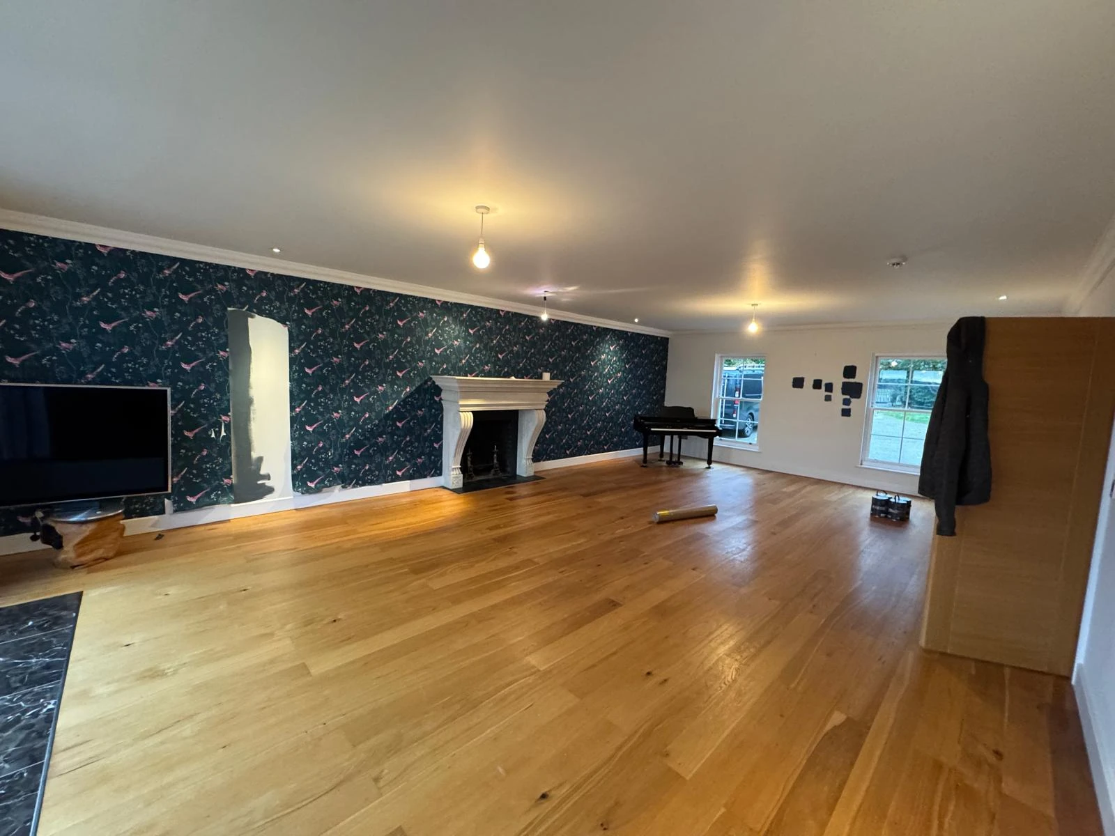 Spacious room with patterned wallpaper, a fireplace, wooden flooring, and a piano. Large windows let in natural light.