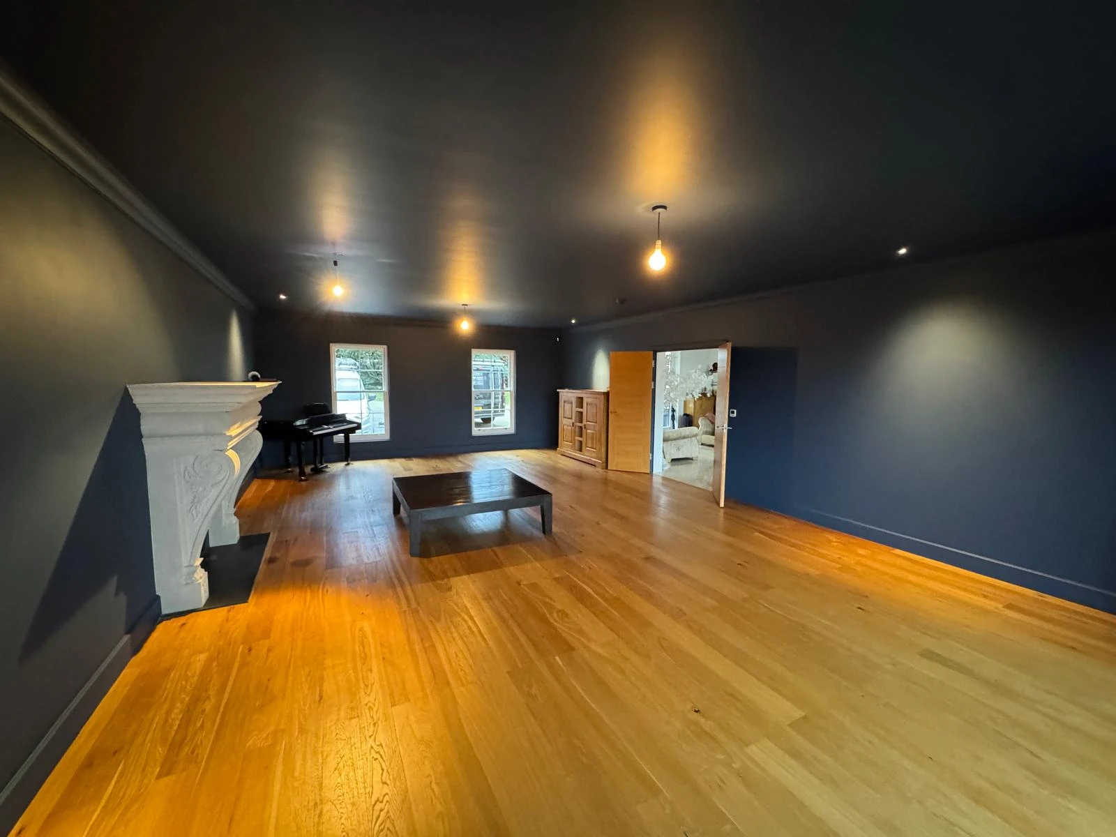 Spacious room with dark blue walls, wooden flooring, a fireplace, a piano, and warm lighting, leading to another area.