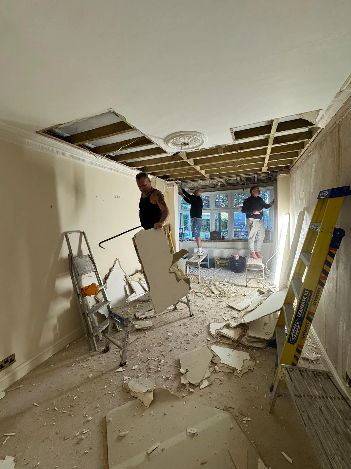 Workers renovating a room with a partially removed ceiling, debris on the floor, and ladders present. Natural light from windows.