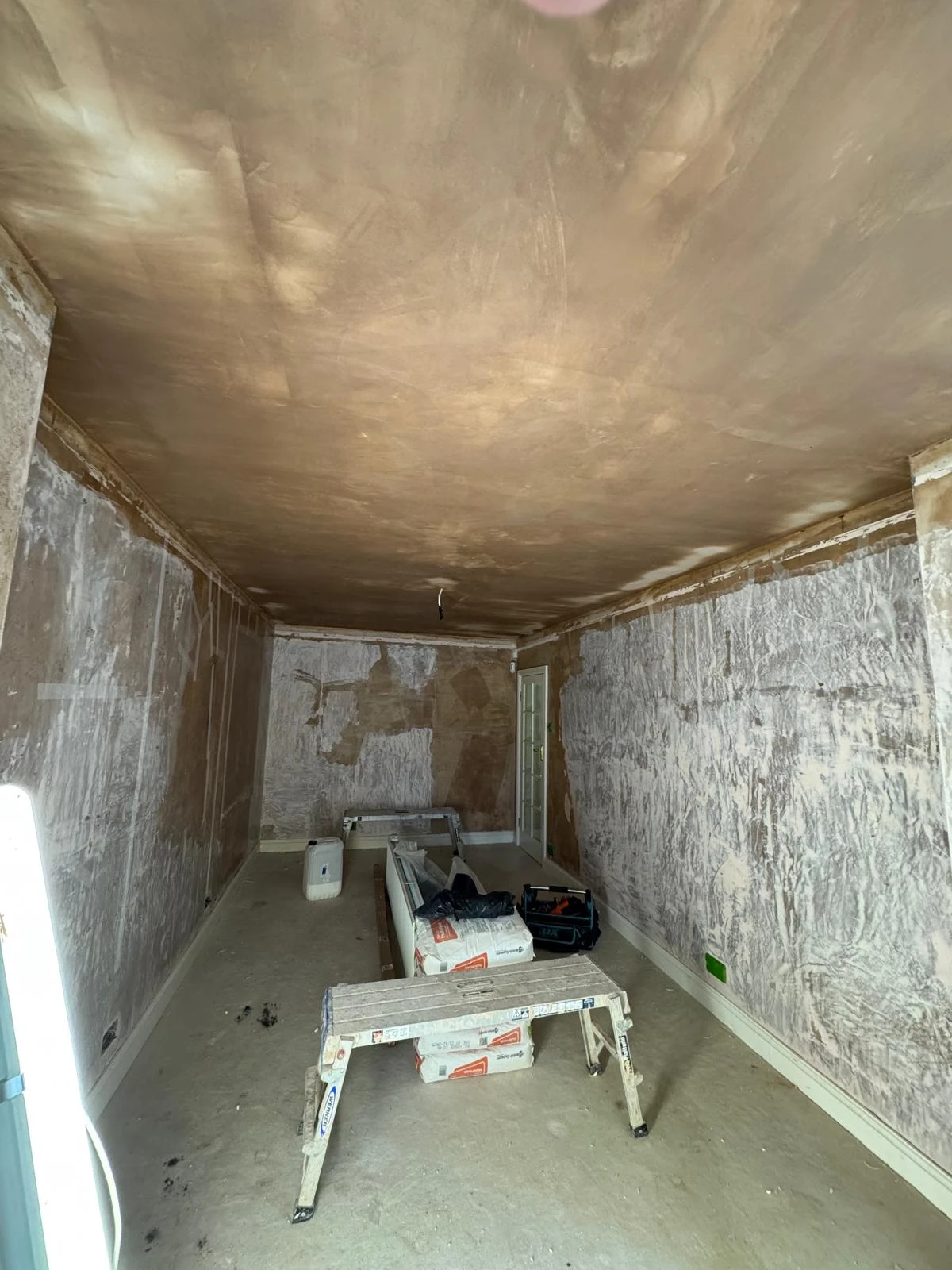 An empty, partially renovated room with messy walls and a ceiling. There are tools and materials scattered on the floor and a ladder.