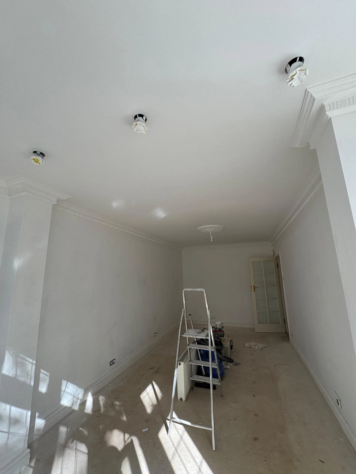 A partially furnished room with a ladder, paint tools, and white walls, featuring ceiling lights and a closed glass door.