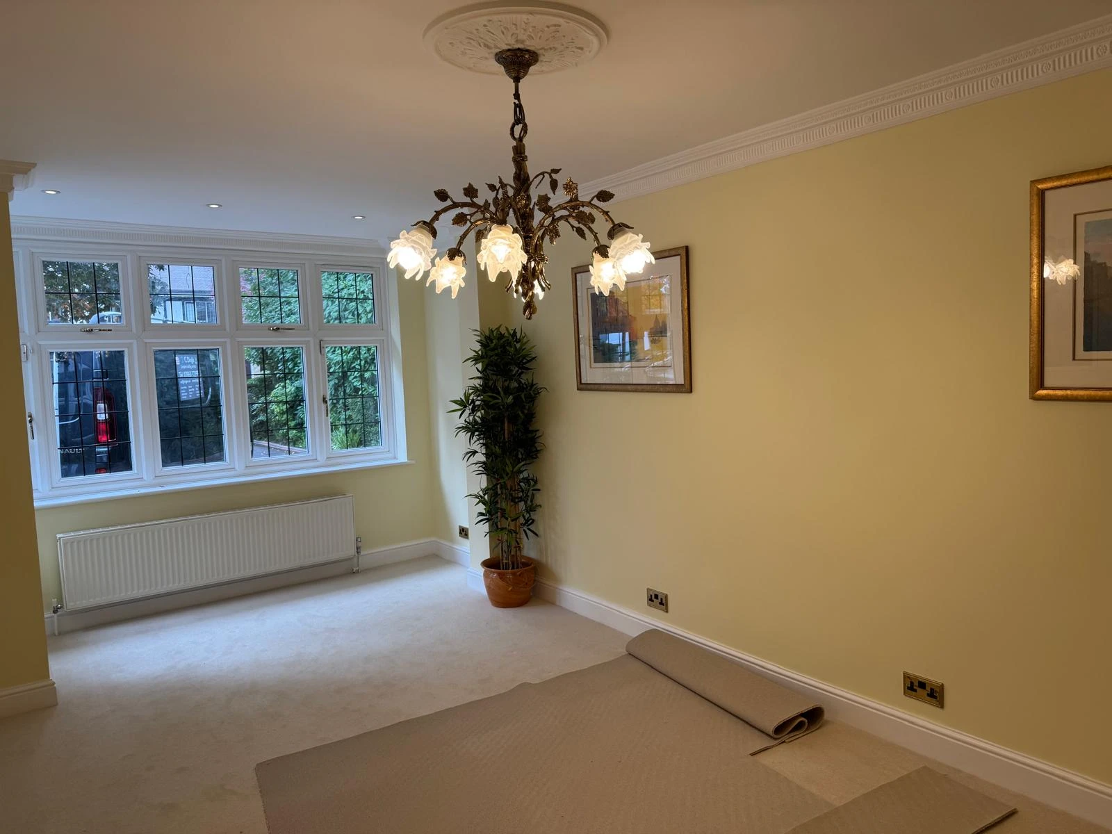 Bright room with yellow walls, large windows, and a decorative chandelier. Includes a potted plant and rolled-up carpet.