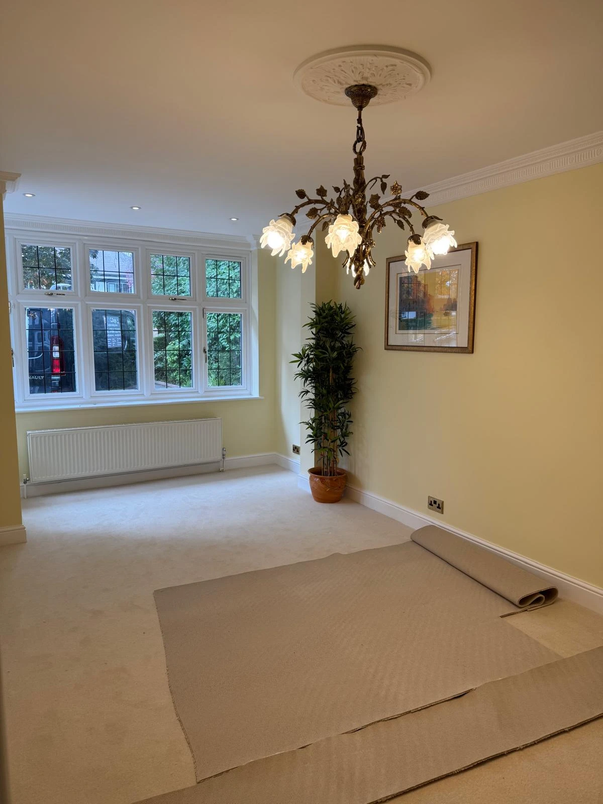 A bright, spacious room with yellow walls, a chandelier, and large windows. A rolled-up carpet lies on the floor next to a potted plant.